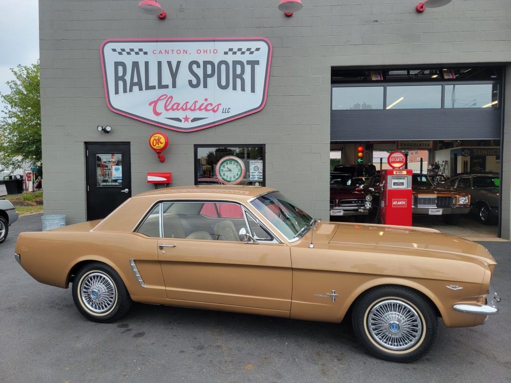 A tan 1965 Ford Mustang is parked in front of a rally sport classics store.