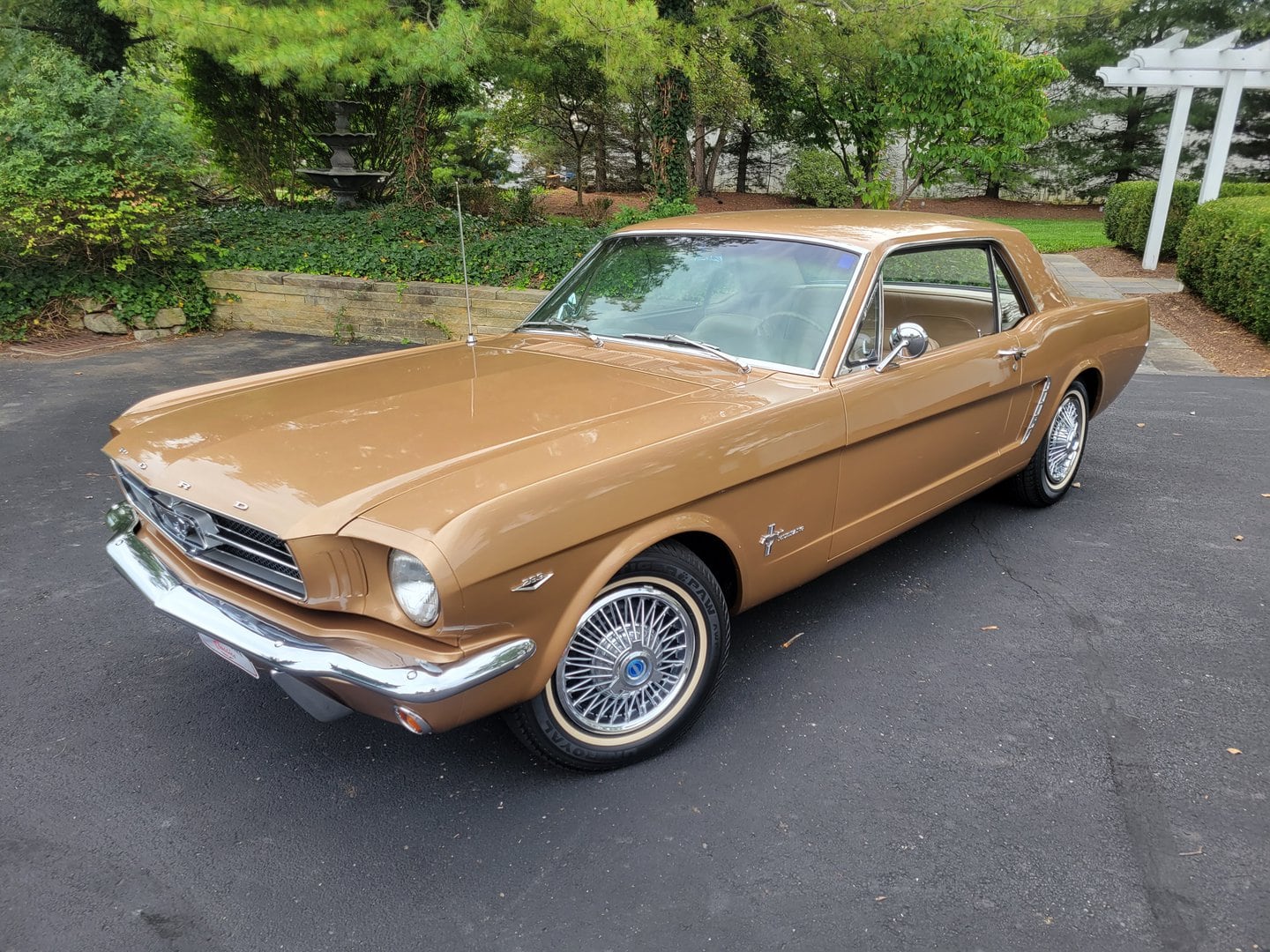 A 1965 tan Ford Mustang is parked in a driveway.