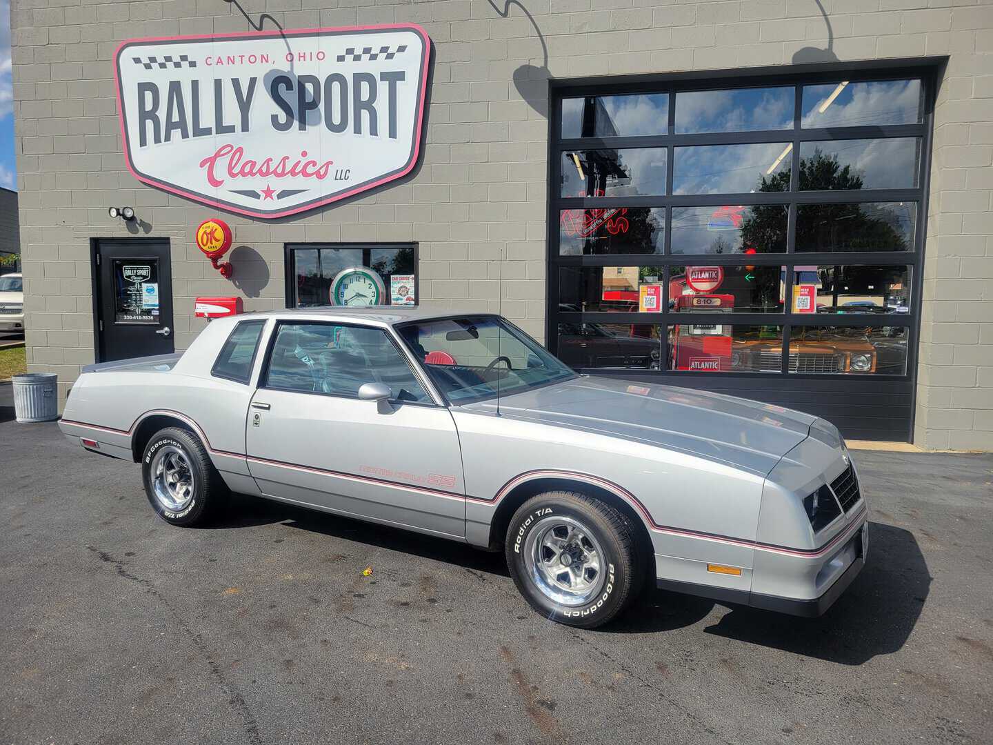 A silver Buick Regal is parked in front of a race car shop featuring a 1985 Monte Carlo SS.
