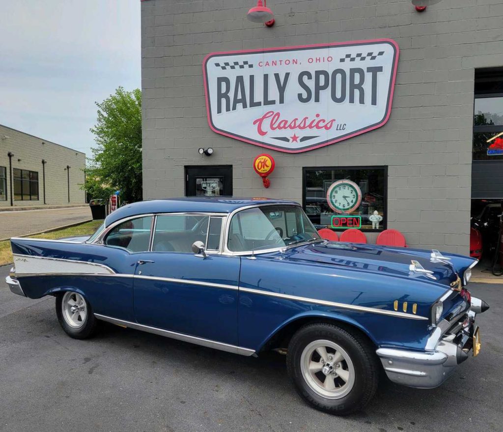 A 1957 Chevrolet Bel Air, A Blue Classic Car, Is Parked In Front Of A Bully Sport Classics Store.