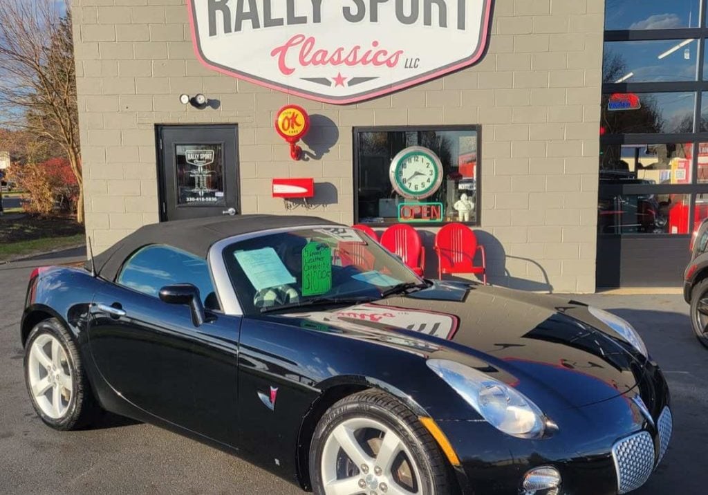 A 2006 Pontiac Solstice, A Black Sports Car, Is Parked In Front Of A Rally Sport.