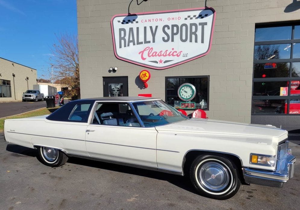 A 1975 White Coupe Deville Cadillac Parked In Front Of A Rally Sport Store.