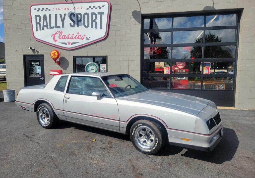 A silver Buick Regal is parked in front of a race car shop featuring a 1985 Monte Carlo SS.
